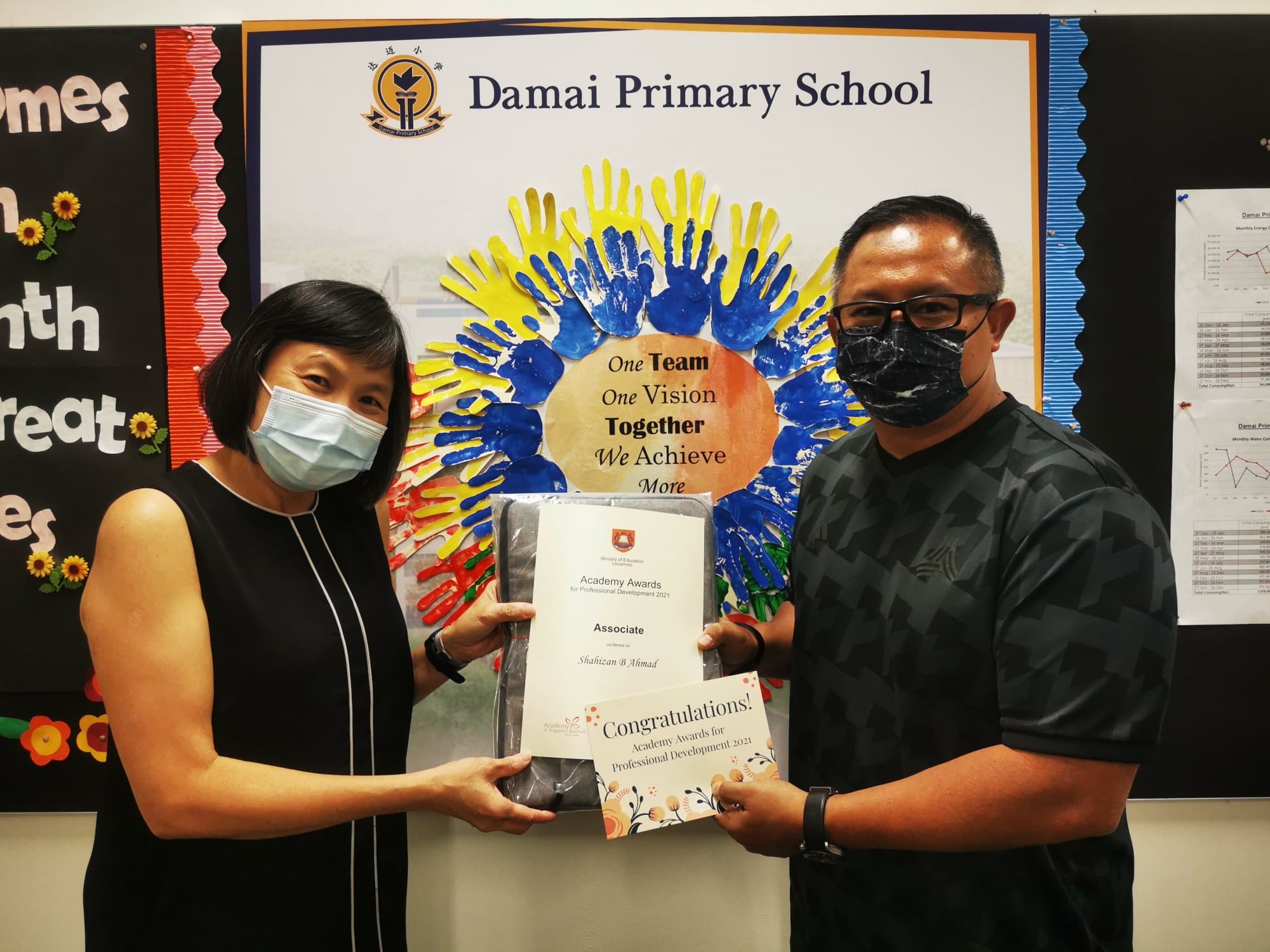 Two masked people hold an Academy Awards certificate at Damai Primary School in front of a colorful handprint display.